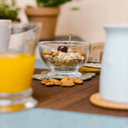 Clear footed bowl holding cereal, nuts and a cherry on a breakfast table
