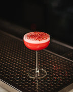 Clear stemmed coupe holding a red drink with a pale foam top on a black bar mat