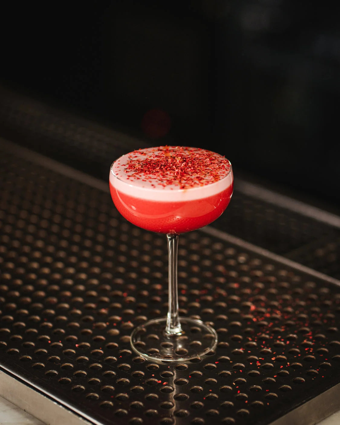 Clear stemmed coupe holding a red drink with a pale foam top on a black bar mat